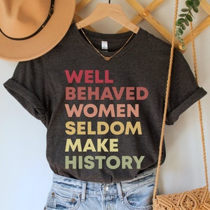 May include: A dark gray t-shirt with the text "WELL BEHAVED WOMEN SELDOM MAKE HISTORY" printed in a gradient of pink, orange, and yellow.