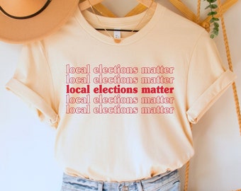 May include: A beige t-shirt with the text "local elections matter" repeated five times in red and white lettering. The shirt is hanging on a wooden hanger with a straw hat and a woven bag.