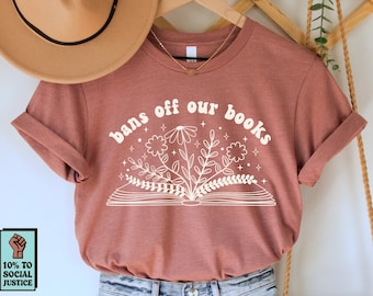May include: A brown t-shirt with the text "bans off our books" in white with a stylized illustration of an open book with flowers and stars. The shirt is hanging on a wooden rack with a brown hat and a woven bag.