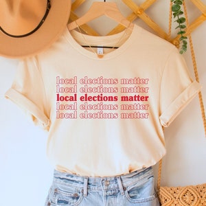 May include: A beige t-shirt with the text "local elections matter" repeated five times in red and white lettering. The shirt is hanging on a wooden hanger with a straw hat and a woven bag.