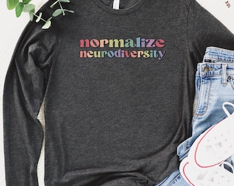 May include: Dark gray long-sleeve shirt with the words "normalize neurodiversity" in a rainbow font. The shirt is paired with blue jeans and white sneakers. A sprig of greenery is in the upper left corner.