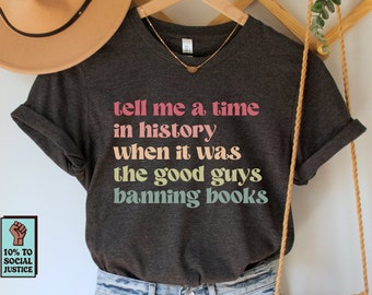 May include: A dark gray t-shirt with the text "tell me a time in history when it was the good guys banning books" in rainbow colors. The shirt is hanging on a wooden hanger with a brown hat and a crocheted bag.