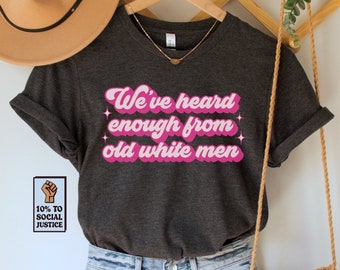 May include: A dark grey t-shirt with the text "We've heard enough from old white men" in pink retro-style lettering. The shirt is hanging on a wooden hanger with a brown woven bag and a brown hat.