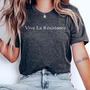 May include: Dark gray t-shirt with the phrase "Vive La Résistance" printed in white. The shirt is paired with light blue jeans. A small tattoo is visible on the arm. A logo indicating a donation to social justice is in the upper right corner.
