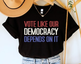 May include: Black t-shirt with the text "VOTE LIKE OUR DEMOCRACY DEPENDS ON IT" in red, white and blue. The shirt is hanging on a wooden hanger with a straw hat and a brown bag.