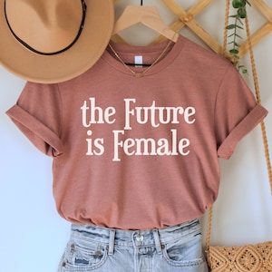 May include: A mauve t-shirt with the text "the Future is Female" in white lettering. The shirt is paired with light blue denim shorts and a brown woven bag with fringe.