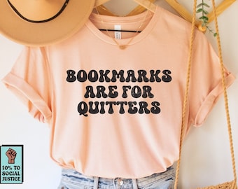 May include: A peach-colored t-shirt with the text "Bookmarks Are For Quitters" in black, paired with denim shorts, a tan hat, and a woven bag. The outfit is displayed on a wooden hanger against a white background.