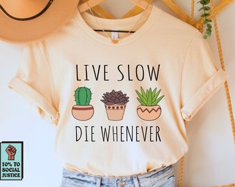 Op de afbeelding: Een crèmekleurig T-shirt met de tekst "LIVE SLOW DIE WHENEVER" en illustraties van cactussen en vetplanten. Het shirt wordt gecombineerd met lichtblauwe denim shorts, een geweven tas en een beige hoed. Een kleine sticker met tekst is ook zichtbaar.