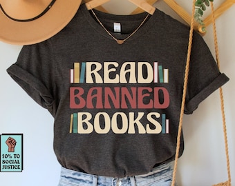 May include: A dark grey t-shirt with the text "READ BANNED BOOKS" in colorful letters. The shirt is hanging on a wooden hanger with a brown hat and a small brown crocheted bag. A small sticker with the text "10% TO SOCIAL JUSTICE" and a raised fist is in the lower left corner.