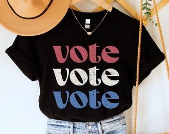 Pode incluir: Uma camiseta preta com a palavra "vote" repetida três vezes em vermelho, branco e azul. A camiseta está pendurada em um cabide de madeira e é combinada com shorts jeans e uma bolsa de palha.