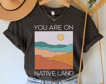 Może przedstawiać: Szary t-shirt z nadrukiem przedstawiającym zachód słońca nad górami i tekstem "You are on Native Land". T-shirt jest połączony z jasnoniebieskimi dżinsowymi szortami i brązową torebką na szydełku.