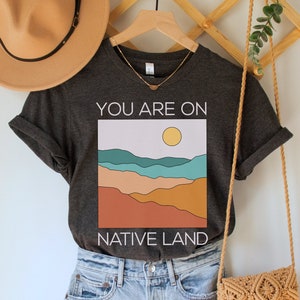 May include: A dark gray t-shirt with a graphic of a sunset over mountains and the text "You are on Native Land". The shirt is paired with light blue denim shorts and a tan crocheted bag.