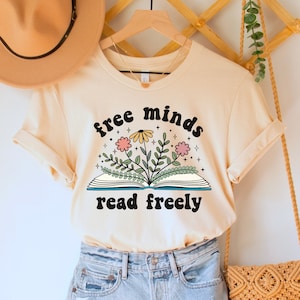 A natural (light beige) unisex crew neck short sleeve t-shirt with black text saying "free minds read freely" around a graphic of a book with colorful flowers and stars coming out of it