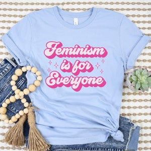 May include: Light blue t-shirt with a pink retro-style graphic that says "Feminism is for everyone" with sparkling stars on either side of the text.