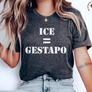 May include: Dark gray t-shirt with the text "ICE = GESTAPO" in white. The shirt has a crew neck and short sleeves. A small logo is visible in the upper right corner. The person is wearing light blue jeans.