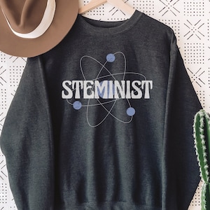 May include: Dark gray sweatshirt with the word "STEMINIST" in light blue and white, featuring a stylized atom graphic. A brown hat with a white band hangs to the left. A cactus in a white pot is on the right.