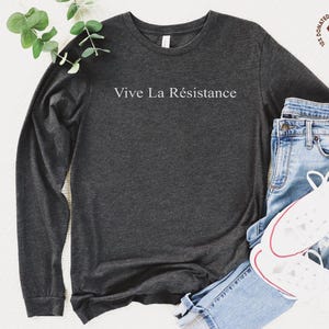 May include: Dark gray long-sleeve shirt with the text "Vive La Résistance" in white. The shirt is paired with blue jeans and white sneakers. A small graphic with the text "10% Donated to Social Justice" is visible.