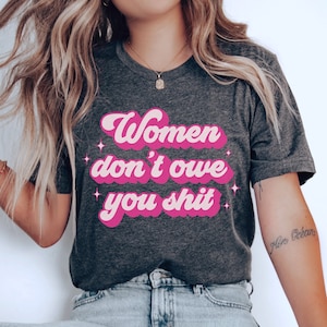 A dark grey heather unisex crew neck t-shirt with the words "women don't owe you shit" in retro pink text with retro pink stars