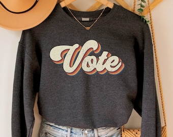 May include: A dark gray sweatshirt with a retro-style "Vote" graphic in rainbow colors. The sweatshirt is hanging on a wooden hanger with a straw hat and a woven bag.