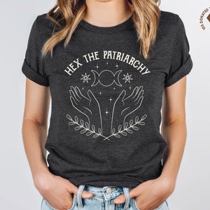 A dark grey heather unisex crew neck short sleeve Bella+Canvas t-shirt that says "hex the patriarchy" in a white uppercase distressed font, above a witchy white graphic with leaves, hands, stars, and a three moon symbol