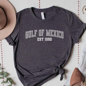 A dark grey heather unisex crew neck short sleeve Bella+Canvas brand t-shirt that says "GULF OF MEXICO" on the chest in white outline college style font with white stripes inside the letters. The text is curved over smaller solid white letters in the same font that say "EST 1550". The graphic has a distressed/vintage look