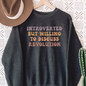 May include: A dark gray sweatshirt with the text "INTROVERTED BUT WILLING TO DISCUSS REVOLUTION" printed in colorful letters.