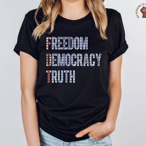 A black unisex crew neck short sleeve Bella+Canvas t-shirt that says "FREEDOM DEMOCRACY TRUTH" on the chest in three lines in blue block text with white stars inside the letters. The first letter of each word in in red with white stars, subtly spelling "FDT" down the shirt