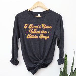 May include: A dark gray long-sleeved t-shirt with the text "I Don't Care What the Bible Says" in a retro yellow and pink font.