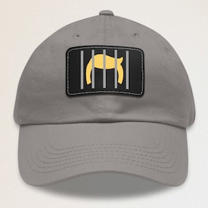 May include: A gray baseball cap with a black patch featuring a yellow silhouette of a person's hair behind bars. The patch has the text "Jail"