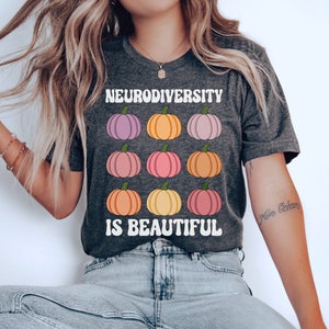 May include: A dark gray t-shirt with the text "Neurodiversity is beautiful" in white letters. The shirt features a pattern of nine colorful pumpkins.