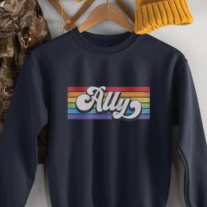 Pu&ograve; includere: Felpa blu navy con un design a righe arcobaleno e la parola "Ally" in un font retr&ograve;.