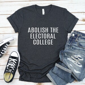 May include: A dark grey t-shirt with the text "Abolish the Electoral College" printed in white on the front. The shirt is laid flat on a white wooden surface with a pair of black sneakers and blue jeans.