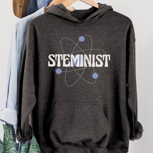 May include: A dark gray hooded sweatshirt with a white graphic of an atom with the word "STEMINIST" printed below it.