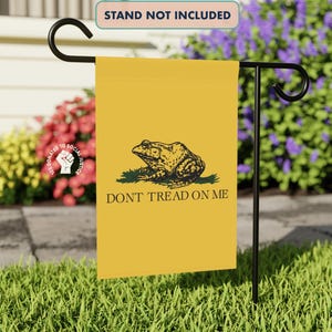 May include: A yellow garden flag featuring a detailed illustration of a frog on a patch of grass, with the words "DON'T TREAD ON ME" below. The flag is displayed on a black metal stand in a garden setting.