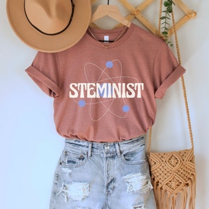 May include: A rust-colored t-shirt with a white atom graphic and the text "STEMINIST" in blue letters. The shirt is hanging on a wooden hanger with a brown hat and a woven bag.