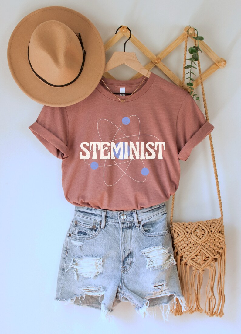 May include: A rust-colored t-shirt with a white atom graphic and the text "STEMINIST" in blue letters. The shirt is hanging on a wooden hanger with a brown hat and a woven bag.