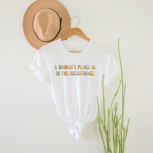 May include: White t-shirt with the text "A woman's place is in the resistance" in colourful, rainbow-like letters.
