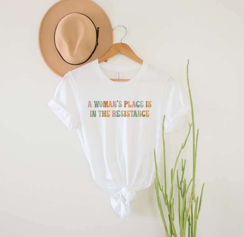 May include: White t-shirt with the text "A woman's place is in the resistance" in colourful, rainbow-like letters.