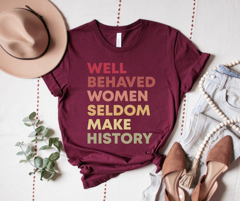 May include: Burgundy t-shirt with the text "WELL BEHAVED WOMEN SELDOM MAKE HISTORY" in a gradient of yellow, orange, and red.