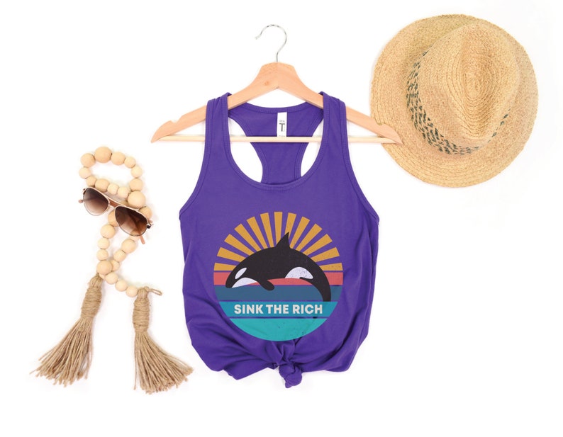 A purple women's racerback tank top that says "sink the rich" in white over a retro graphic of an orca whale in the ocean