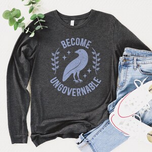 A dark grey heather unisex long sleeve crew neck t-shirt that says  "BECOME UNGOVERNABLE" on the chest in block text around an image of a crow, leaves, and retro stars. The whole graphic and text are light blue and have a distressed/vintage look.