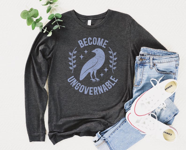 A dark grey heather unisex long sleeve crew neck t-shirt that says  "BECOME UNGOVERNABLE" on the chest in block text around an image of a crow, leaves, and retro stars. The whole graphic and text are light blue and have a distressed/vintage look.