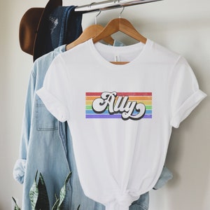 May include: White t-shirt with a rainbow stripe design and the word "Ally" in a retro font.