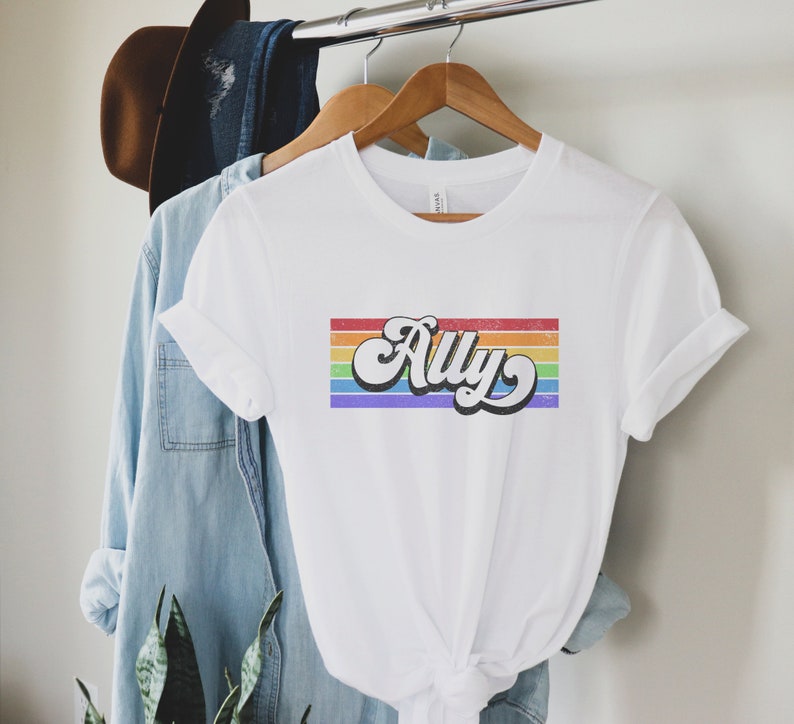 May include: White t-shirt with a rainbow stripe design and the word "Ally" in a retro font.