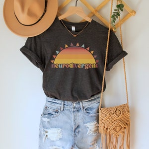 May include: A dark gray t-shirt with a sunset graphic and the text "neurodivergent" in rainbow colors. The shirt is on a hanger with a straw hat and a woven bag.