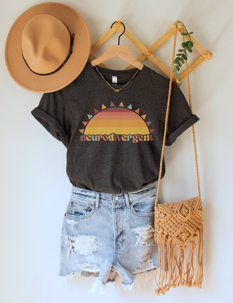 May include: A dark gray t-shirt with a sunset graphic and the text "neurodivergent" in rainbow colors. The shirt is on a hanger with a straw hat and a woven bag.