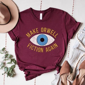 A maroon unisex crew neck Bella Canvas brand t-shirt that says "MAKE ORWELL FICTION AGAIN" in orange text with a black outline, around an image of a blue eye. The graphic has a distressed vintaged look.