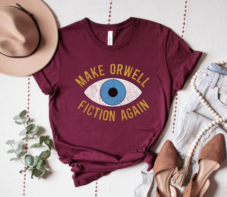 A maroon unisex crew neck Bella Canvas brand t-shirt that says "MAKE ORWELL FICTION AGAIN" in orange text with a black outline, around an image of a blue eye. The graphic has a distressed vintaged look.