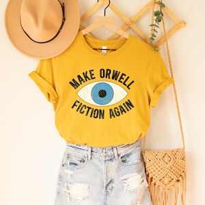 A mustard yellow unisex crew neck Bella Canvas brand t-shirt that says "MAKE ORWELL FICTION AGAIN" in black text, around an image of a blue eye. The graphic has a distressed vintaged look.