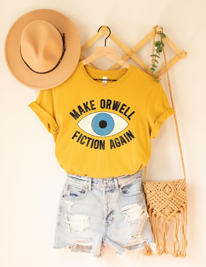 A mustard yellow unisex crew neck Bella Canvas brand t-shirt that says "MAKE ORWELL FICTION AGAIN" in black text, around an image of a blue eye. The graphic has a distressed vintaged look.